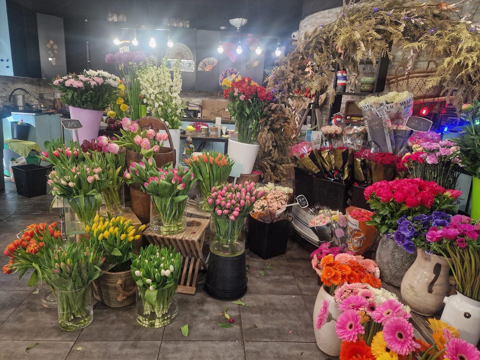 L’intérieur de la boutique : tulipes, roses, pivoines, gerberas et anémones en seaux, fleurs séchées sous les guirlandes
